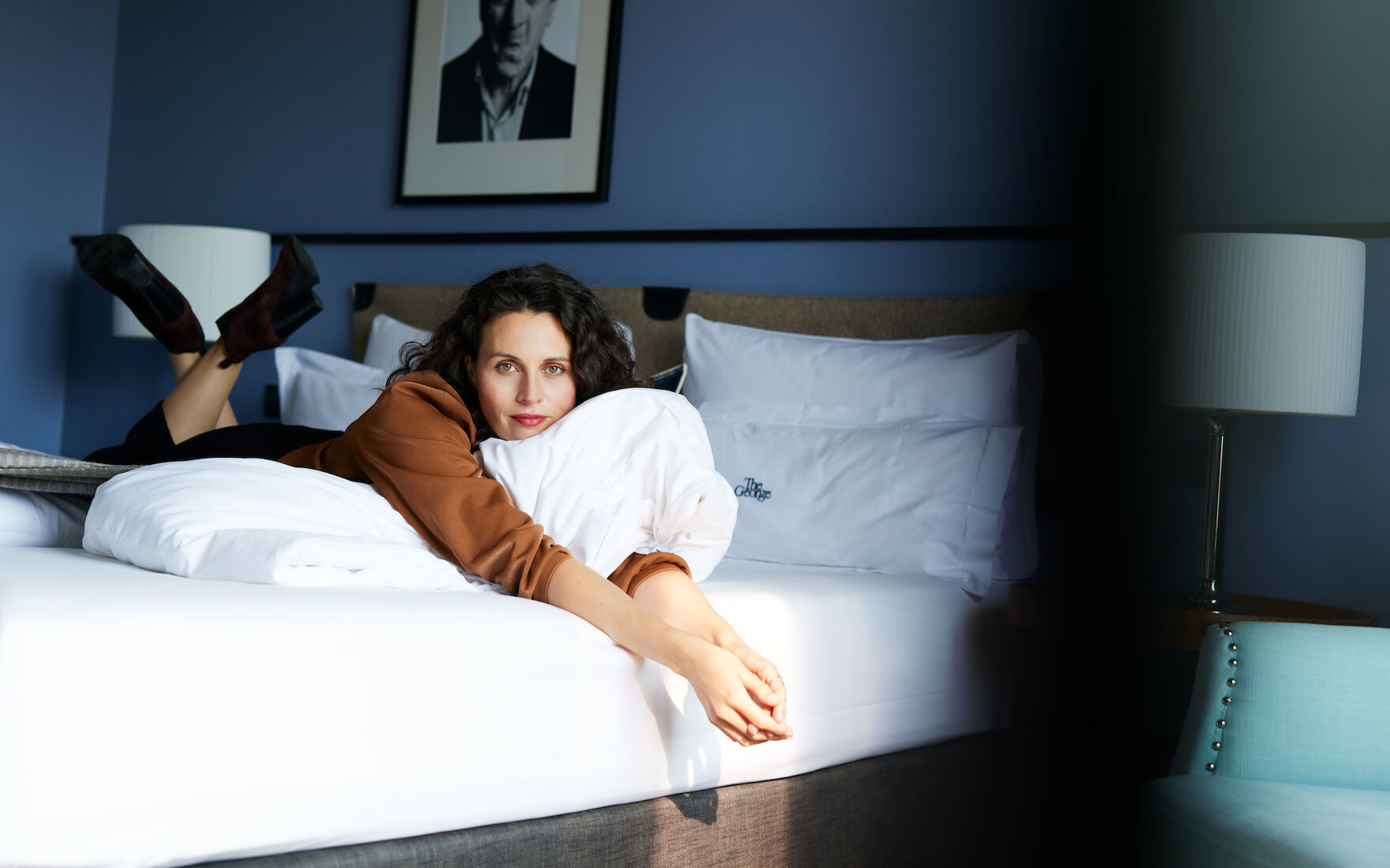 Woman lying in bed in M-Room with blue wallpaper and wall art behind