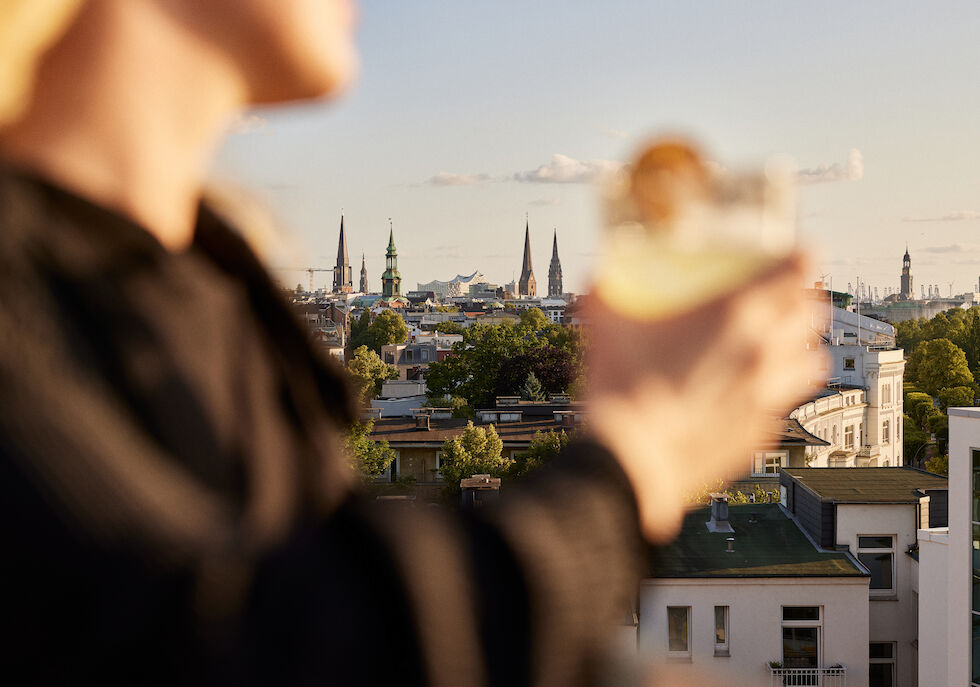 A blurred figure holding a drink in the foreground, with the Hamburg skyline in the background